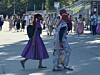 Zwei kostümierte Frauen gehen bei einer belebten Veranstaltung im Freien an einer Menschenmenge vorbei.