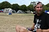 Bärtiger Mann mit Stirnband sitzt vor Zelten auf einer Wiese.