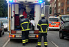 Zwei Feuerwehrkräfte stehen hinter einem Rettungsfahrzeug auf einer innerstädtischen Straße.