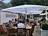 Gäste essen und trinken an Holztischen auf einer begrünten Restaurantterrasse unter Sonnenschirmen.