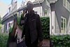 Unkenntliche Person in dunkler Kleidung steht vor Wohnhäusern und Hecken.