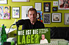 Mann spricht in einem Raum mit Bildern vor einem Mikrofon und einem grünen Schild „Wie ist die Lage?“