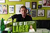 Mann spricht in einem Raum mit Bildern vor einem Mikrofon und einem grünen Schild „Wie ist die Lage?“