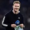 Lächelnder Fußballer im schwarzen VfL-Wolfsburg-Hoodie hält ein Mikrofon und ein graues Kleidungsstück.