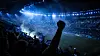 Fans feiern nachts im voll besetzten Stadion, eine erhobene Faust vor dem blau beleuchteten Spielfeld.