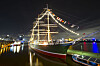 Beleuchtetes historisches Segelschiff liegt nachts im Hafen; Lichter spiegeln sich im Wasser.