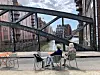 Drei Menschen sitzen auf Stühlen unter einer Stahlbrücke in einer urbanen Straße.