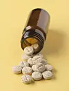 Gesprenkelte runde Tabletten liegen vor einer offenen braunen Glasflasche auf gelbem Untergrund.