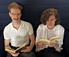 Zwei junge Menschen lesen nebeneinander in aufgeschlagenen Büchern vor dunklem Hintergrund.