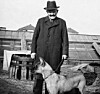 Bärtiger Mann mit Hut und Mantel steht neben einem Deutschen Schäferhund vor Gewächshäusern.