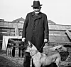 Bärtiger Mann mit Hut und Mantel steht neben einem Deutschen Schäferhund vor Gewächshäusern.
