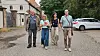 Vier Erwachsene gehen auf einem Schotterweg an historischen Gebäuden und geparkten Autos vorbei.