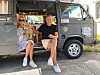 Zwei Erwachsene sitzen vor einem grauen Campingbus in der Sonne.