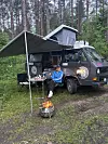 Person sitzt vor einem Expeditionsfahrzeug im Wald, während davor ein kleines Lagerfeuer brennt.