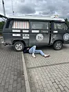 Person liegt teilweise unter einem dunkelgrünen Kleinbus auf einem gepflasterten Parkplatz.