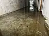 Wasser überflutet einen langen Kellerflur mit sichtbaren Leitungen und einer Tür im Hintergrund.