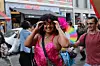 Person in pinkem Paillettentop posiert bei einer farbenfrohen Pride-Parade auf einer belebten Straße.