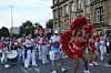 Tänzer in roten und weißen Kostümen führen eine Parade mit Trommeln durch eine belebte Innenstadtstraße an.