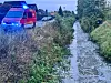 Rotes Einsatzfahrzeug mit Blaulicht steht neben einem bewachsenen Bachlauf an einer Straße.
