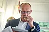Bundeskanzler Friedrich Merz isst ein Fischbrötchen und hält Unterlagen in einem hellen Innenraum.