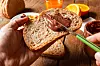 Hand bestreicht eine Brotscheibe mit Schokoladenaufstrich; daneben stehen Orangensaft und Orangen.