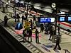 Fahrgäste warten auf einem U-Bahn-Bahnsteig unter digitalen Anzeigetafeln.