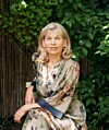 Frau in gemustertem Kleid sitzt vor einem Holzzaun in einem grünen Garten.