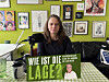 Frau hält in einem bunt dekorierten Raum ein grünes Schild mit der Aufschrift „Wie ist die Lage?“