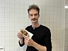 Mann mit Schnurrbart hält in einer gekachelten Küche ein eingewickeltes Gericht.
