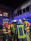 Feuerwehrleute in Schutzkleidung blicken nachts auf eine Einsatzstelle vor einem blau beleuchteten Gebäude.