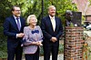 Drei festlich gekleidete Menschen stehen neben einer Büste in einem begrünten Garten.