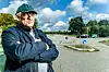 Mann mit grüner Kappe und dunkler Jacke steht mit verschränkten Armen auf einem Parkplatz.