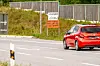 Roter Pkw fährt auf einer Straße an einem handbeschriebenen Hinweis und einem Zaun vorbei.
