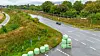 Grüne Rundballen liegen an einer kurvigen Straße; im Hintergrund fährt ein Auto durch eine bewaldete Landschaft.