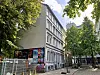 Bäume säumen eine Straße mit Altbauten, Plakatwänden, Bauzaun und Fahrrädern.