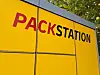Gelbe DHL-Packstation in Freiburg mit großem PACKSTATION-Schriftzug unter Bäumen.