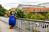 Lächelndes Maskottchen in blauem Trikot winkt auf einer Fußgängerbrücke vor einem Backsteingebäude.