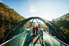 Zwei Menschen gehen über eine Fußgängerbrücke vor bewaldeten Berghängen im Sonnenlicht.