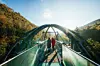 Zwei Menschen gehen über eine Fußgängerbrücke vor bewaldeten Berghängen im Sonnenlicht.
