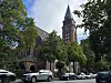 Historische Backsteinkirche mit spitzem Turm, Bäumen und Autos an einer Stadtstraße