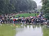 Große Menschenmenge steht an einem von Bäumen gesäumten Teich in einer Parkanlage.