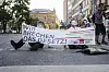 Zwei Personen der Letzten Generationen blockieren eine Straße. Jeweils eine Hand ist an die Fahrbahn geklebt, mit der anderen halten sie ein Banner „Wir brechen das Gesetz!“, mit den Logos der Regierungsparteien. Ein Teilnehmer trägt eine Olaf-Scholz-Maske, der andere eine Robert-Habeck-Maske.