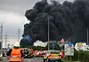 Einsatzfahrzeuge der Feuerwehr stehen unweit einer Zufahrt zum Chempark über dem eine dunkle Rauchwolke aufsteigt.