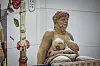 Eine sitzende, nackte Frau mit hängenden Brüsten, Schwabbelbauch und einem Gesicht, das stark an Angela Merkel erinnert