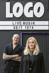 Chris August und Lea Goltz stehen in schwarzer Kleidung vor dem Logo