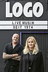 Chris August und Lea Goltz stehen in schwarzer Kleidung vor dem Logo