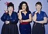 Drei Frauen in blauen Kleidern mit roten Details lachen in die Kamera
