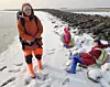 Eine Frau und ihre zwei Kinder stehen und sitzen am Wattenmeer. Die Gesichter der Kinder sind gepixelt.